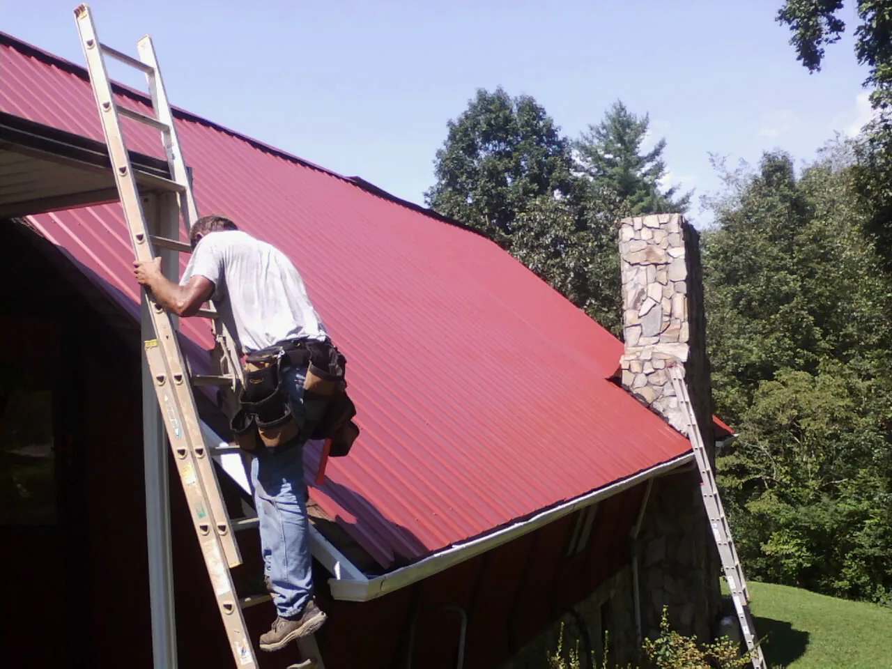 Roofer on a ladder beside a red metal roof and stone chimney — Soffit & Fascia Repair by Roofing Brothers in Beechmont